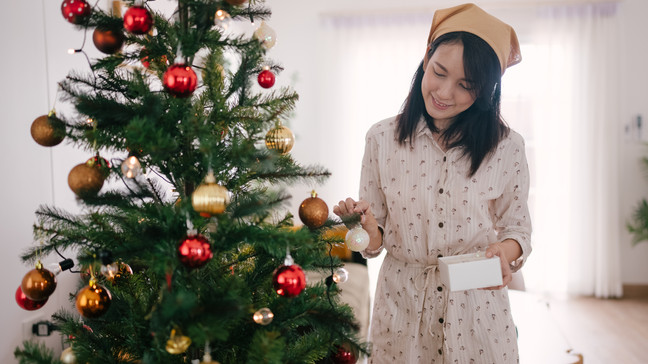 Woman preparing and hanging decorations on Christmas tree at home. (Credit: Getty Images)